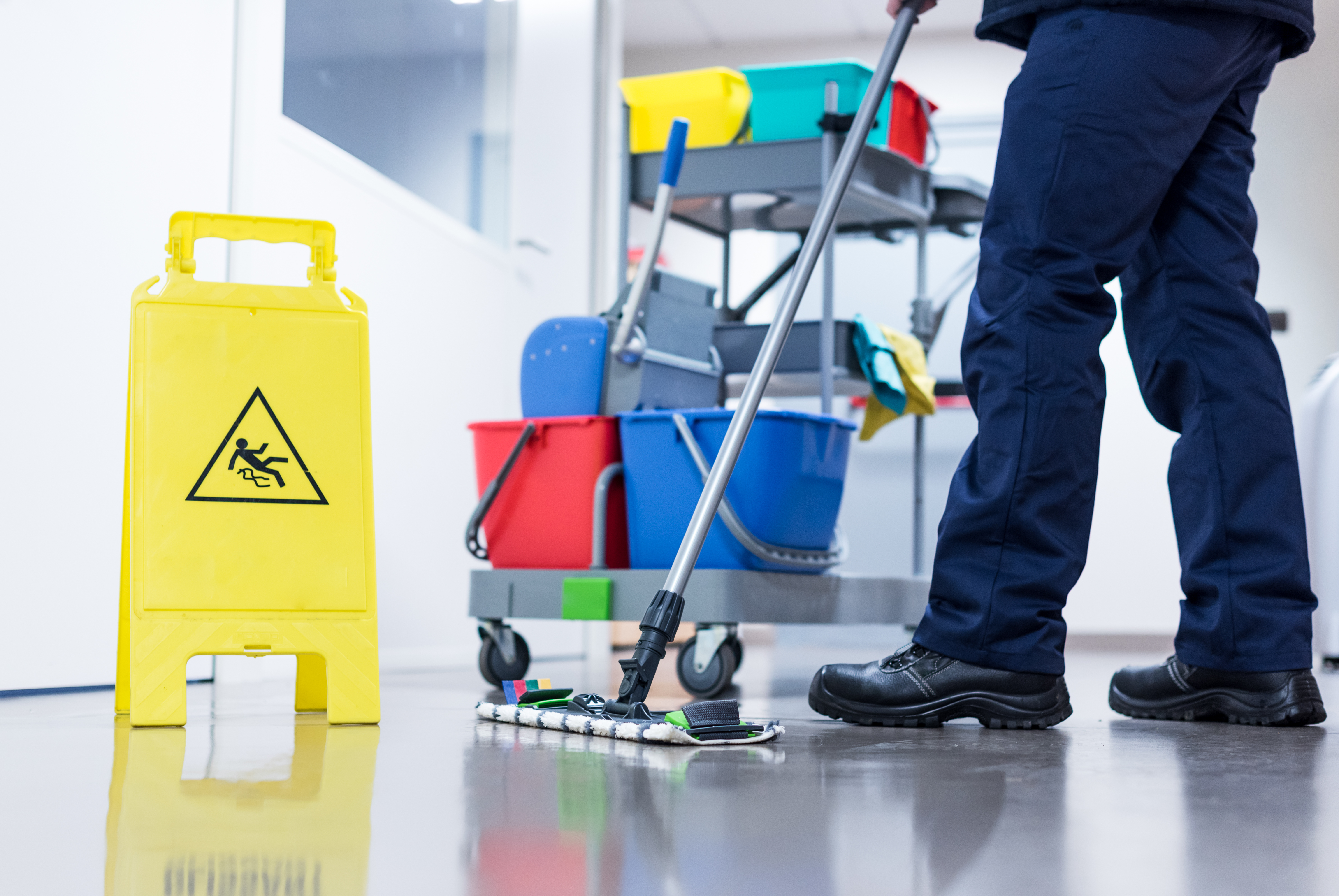 Janitor mopping floor with wet mop near yellow wet floor caution sign and cleaning supply cart